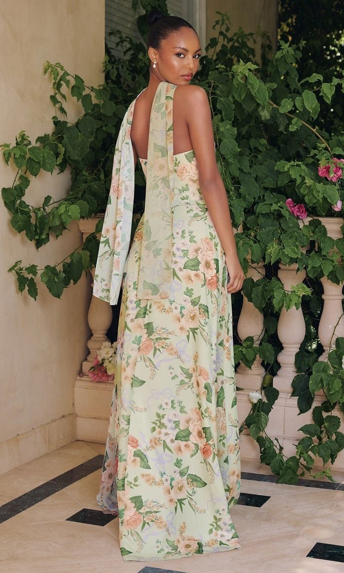 A woman stands outdoors in the Amoris T717 Strapless Long Print Prom Dress, featuring a floral pattern and tie-back detail, looking over her shoulder amid green foliage and flowers.